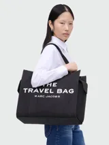 Bolso tote de lona color negro, con asas de mano y correa de hombro. Estampado frontal con el texto "THE TRAVEL BAG MARC JACOBS" en color blanco.