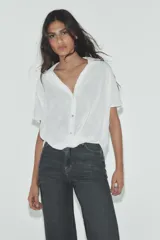 Camisa color gris claro con cuello solapa y escote pico, de manga corta. Presenta un nudo en la parte delantera y cierre frontal con botones.