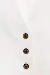 Chaleco blanco con cuello en V, sin mangas, con cierre frontal de botones marrones a contraste y bolsillos simulados en la parte delantera.