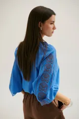 Camisa blanca de manga larga con bordados marrones en hombros y mangas, cuello en V y cierre frontal con botones.