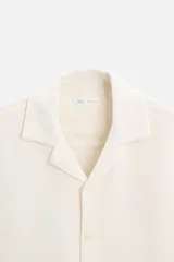Camisa color crudo de corte regular fit, confeccionada en tejido con textura. Tiene cuello bowling, manga corta, bajo con aberturas laterales y cierre frontal con botones.