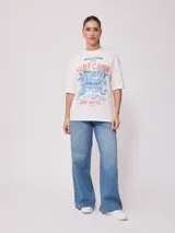 Remera blanca de algodón con estampado frontal de estilo playero con texto "Welcome to the Surf Camp" y dibujo de una casa de playa con palmeras y tablas de surf.