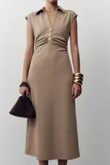 Vestido midi color beige con cuello polo, escote en pico, manga sisa caída y detalle de drapeado lateral. Cierre frontal con botones metálicos en relieve.