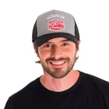 Gorro trucker de hombre, con visera plana, frente gris con logo bordado de Caterpillar y laterales de rejilla negra.
