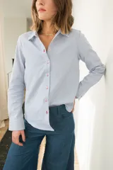 Camisa celeste a rayas verticales blancas, de corte clásico con cuello camisero, mangas largas y cierre frontal con botones rojos. Presenta costuras en contraste y un corazón bordado en la espalda.