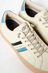 Zapatillas deportivas de cuero sintético color blanco hueso, con suela marrón claro y detalles en azul y negro.