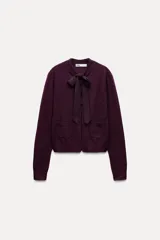 Chaqueta color borgoña de tejido de punto con lana y alpaca. Tiene cuello redondo con lazo de satén, manga larga y cierre frontal con botones. Presenta dos bolsillos de parche en el frente.