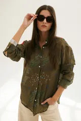 Camisa beige de manga larga con cuello clásico y cierre frontal con botones. Presenta bordados con motivos florales y geométricos en tonos azules, rojos y verdes.