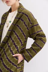 Kimono largo verde oliva con estampado batik en tonos morados. Tiene mangas largas, bolsillos laterales y se ata a la cintura con un cinturón blanco y negro.