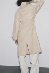 Abrigo largo color beige confeccionado en hilatura con lana. Tiene cuello subido, manga larga con trabillas, bolsillos laterales y cinturón ajustable con lazada. El cierre frontal es cruzado con botones.