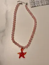 Collar de cuentas de piedras naturales rosadas con un dije de estrella de mar de cristal rojo con puntos blancos y bordes amarillos. Tiene cierre de mosquetón y cadena de extensión plateada.