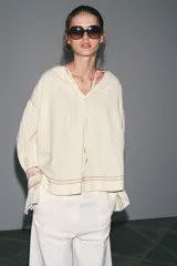 Camisa color crudo de cuello redondo con escote pico y lazada. Manga larga acabada en volantes. Detalle de bordados combinados a contraste y puntilla a tono.