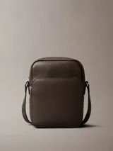 Cartera bandolera Calvin Klein de piel sintética texturizada, color marrón oscuro, con silueta estructurada y correa ajustable. Presenta un bolsillo exterior frontal y un compartimento principal con cierre de cremallera.