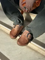 Zuecos de cuero color marrón claro con hebilla metálica y detalle de ojales.