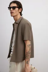 Camisa marrón de punto con textura, corte regular, cuello inglés, tapeta lisa, canesú en la espalda, bajo recto con aberturas laterales y manga corta.
