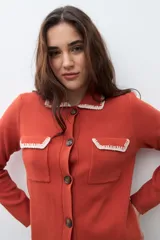 Chaqueta de tejido color terracota/lacre, con cuello y bordes de bolsillo en blanco con textura de punto. Posee cierre frontal de botones y manga larga.