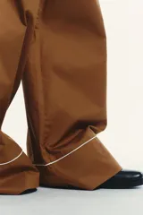 Pantalón marrón de popelina con corte palazzo, tiro medio, cintura elástica ajustable con cordones, bolsillos laterales y falsos bolsillos de vivo con botones en la espalda. Detalle de vivos blancos a contraste en el ruedo.