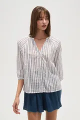 Camisa blanca de algodón con rayas verticales en tono beige/tostado y detalles de bordado tipo ojal a lo largo de las rayas. Presenta cuello mao con abertura en V y botones frontales, y mangas 3/4 con ligero volumen.