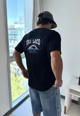 Remera negra de corte holgado con estampado gráfico en la espalda que dice "TAS LOCO" en letras rosas y celestes, con una ilustración de una ola y el texto "SUNNY WITH A CHANCE OF STOKE".