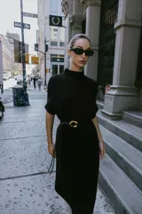 Vestido negro de tejido de punto, corte midi y silueta holgada, con cuello alto acanalado y mangas cortas tipo murciélago. Se ajusta a la cintura con un cinturón fino de efecto piel negro con hebilla dorada en forma de C.