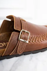 Zueco de cuero color marrón con diseño trenzado en la puntera, hebilla metálica ajustable y plantilla de corcho.