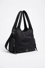 Bolso tipo shopper de nylon negro, con estructura blanda y acabado ligeramente arrugado. Cuenta con doble asa de mano y una correa ajustable y extraíble para llevar al hombro o cruzado. Presenta bolsillos exteriores con cierre de cremallera a cada lado y el logo metálico de Bimba y Lola en la parte inferior frontal.