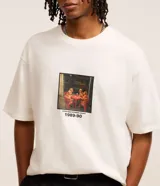 Remera de corte boxy color crudo, con cuello redondo y mangas cortas. Estampa frontal con dibujo de perros jugando a las cartas y texto 'underappreciated crew 1989-90'.