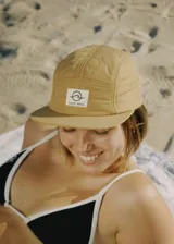 Gorra de cinco paneles color camel con logo estampado en etiqueta blanca en el frente.