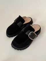 Zueco tipo mule negro, confeccionado en cuero vacuno, con puntera cuadrada y suela track gruesa. Presenta una faja ancha con hebilla metálica plateada sobre el empeine.