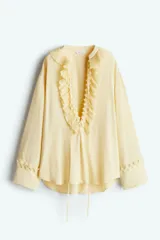 Túnica color amarillo pálido de corte holgado, con cuello mao y abertura frontal con volados plisados. Mangas largas con puños anchos con volados y botones. Bajo recto.