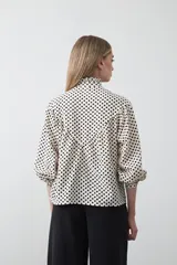 Camisa de viscosa blanca con estampa de lunares negros. Presenta cuello alto con volado y botonadura frontal, mangas largas con puños abullonados y frunces bajo el busto.