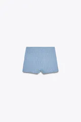 Shorts de tejido con textura color celeste, de tiro alto y cintura elástica. Presenta falsos bolsillos con solapa y botón metálico en el delantero.