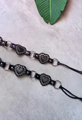 Cinturón de cuero negro con apliques de corazones metálicos en relieve, rodeados de tachas redondas. El cinturón se une mediante argollas metálicas y termina en tiras de cuero para atar.