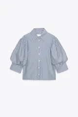 Camisa de algodón a rayas verticales azules y blancas, con cuello solapa y mangas cortas abullonadas. Cierre frontal con botones.