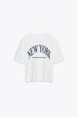 Camiseta blanca de algodón con cuello redondo, corte oversize y mangas cortas. Estampado frontal con la frase "New York" en azul.