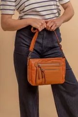 Bandolera de simil cuero color marrón topo, con textura ligeramente desgastada y detalles en nobuck. Presenta múltiples bolsillos frontales con cierres metálicos y tiradores de cuero. Correa ajustable para llevar al hombro o cruzada.