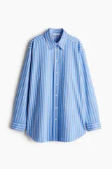 Camisa oversized de popelina de algodón a rayas verticales celestes, blancas y rojas. Tiene cuello clásico, cierre frontal con botones, hombros caídos y mangas largas con puños abotonados. El bajo es redondeado.