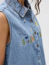 Camisa sin mangas de denim liviano color camel, con cuello camisero y botones frontales. Presenta bordados temáticos marinos en el pecho, incluyendo un sol, olas, una gaviota y vida marina.