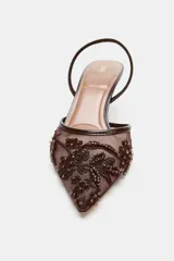 Zapato de tacón alto, destalonado, con punta fina. El cuerpo del zapato es de malla (mesh) color marrón oscuro, con apliques bordados y brillos en tonos similares. Posee una tira trasera elástica para ajuste.