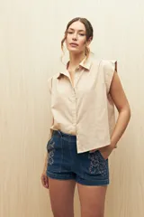 Camisa color beige, de corte recto, con cuello clásico, mangas cortas con volados en los hombros y cierre frontal con botones.