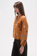 Blusa de algodón color tostado con bordados geométricos en blanco y azul marino, cuello mao con escote en V y mangas largas abullonadas con puños elásticos.