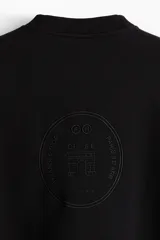 Buzo negro de tejido pesado con interior cepillado suave, cuello redondo y corte boxy. Presenta un diseño estampado en relieve tono sobre tono en el pecho.