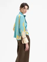 Camisa de manga larga color celeste con estampado de lunas amarillas y cerezas rojas. Presenta cuello con lazo anudado y ribetes en azul marino en el cuello, puños y cartera frontal.