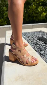 Sandalia de cuero color caramelo con taco ancho de madera. Presenta un diseño de tiras entrecruzadas con recortes geométricos en la parte del empeine y una tira con hebilla metálica plateada en el tobillo.