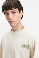 Remera color crema de manga corta con calce boxy, cuello redondo y logo estampado en el pecho. El modelo mide 1,83 y usa talle M.