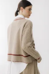 Sweater beige con cuello en V y mangas largas con diseño trenzado. Tiene dos bolsillos frontales con bordados de tréboles rojos y ribetes rojos en cuello, puños y ruedo.