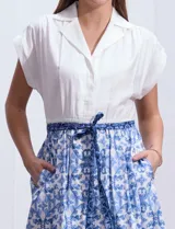 Vestido midi camisero de algodón con estampado floral en tonos azules y blancos. Tiene cuello solapado, cierre frontal con botones, manga corta, cintura elastizada con lazo de ajuste y bolsillos laterales.