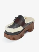 Zapato tipo mule con plataforma track de color marrón y capellada de cuero negro. Cuenta con forro de lana de oveja (borreguito) visible en los bordes e interior, y una correa de cuero marrón con hebilla metálica sobre el empeine.