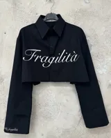 Camisa negra de algodón, corte crop, con cuello clásico, cierre frontal con botones y mangas largas con puños abotonados. Estampado con la palabra "Fragilità" en color blanco en el frente y en la manga izquierda.