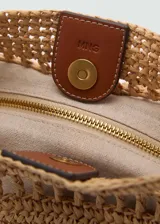 Bolso tipo shopper de paja trenzada color beige con aplique de cuero marrón en el frente. Tiene cierre con botón magnético, bolsillo interior, detalles calados y asa de hombro ajustable con hebillas.